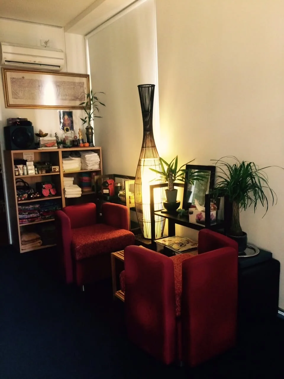 Reception nook at Wanvilai — two red armchairs, woven-rattan floor lamp, shelves of towels and Thai shrine objects